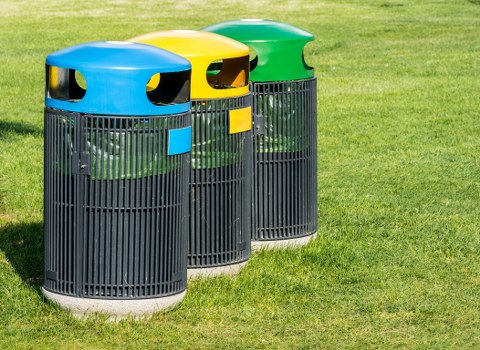 Three cylindrical outdoor rubbish bins positioned side by side on a grassy area, with the foreground bin featuring a blue lid, the middle bin with a yellow lid, and the rightmost bin with a green lid. Each bin is made of black plastic with vertical slats for ventilation and air circulation, and they have smooth, rounded edges and a glossy finish on their lids. The bins are arranged on a paved surface adjacent to a lush green lawn, which extends into the background. The scene appears to be in a public or commercial outdoor space, possibly near a business premises or community area, aligning with the rubbish collection services offered by Commercial Waste Pimlico. Natural sunlight highlights the colours and textures, creating slight shadows around the bases of the bins and reinforcing the clean, orderly appearance of the waste management setup.