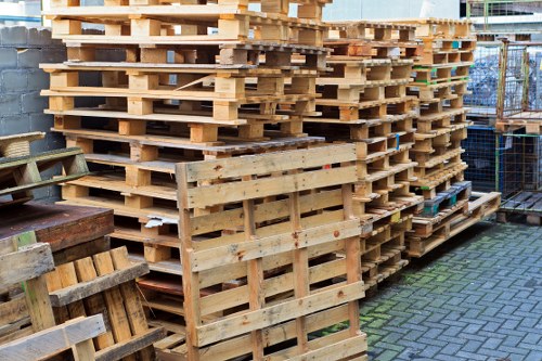 A large stack of untreated wooden pallets arranged outdoors on a paved surface, with some pallets leaning against each other and others stacked neatly in multiple layers. The pallets are made of light-colored timber with visible grain and surface textures, and they vary in condition, with some showing minor wear or weathering. Surrounding the pallets, there are a few additional smaller pallets and wooden crates, indicating a warehouse or industrial yard environment. In the background, there is a chain-link fence and an urban setting, suggesting the area is part of a commercial waste or materials storage site, possibly in Pimlico. The lighting appears natural, with diffuse daylight illuminating the scene, suitable for a professional waste management or rubbish removal context, as handled by companies like Commercial Waste Pimlico.