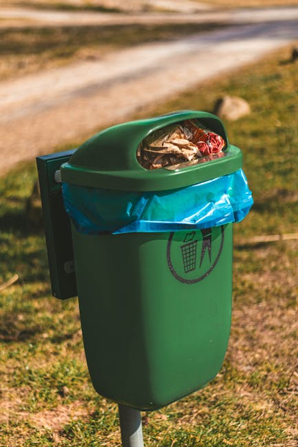 A discarded clear plastic water bottle with a white and blue screw cap lies on a patch of green grass, with small blades and tiny plants visible surrounding it. The bottle appears crushed and empty, w