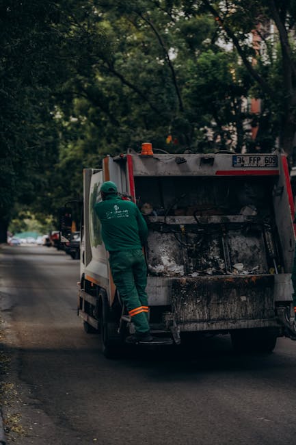 A sanitation worker in a green uniform and protective helmet is seen standing beside the rear of a garbage collection truck on a quiet street lined with trees. The truck, primarily white with red and 