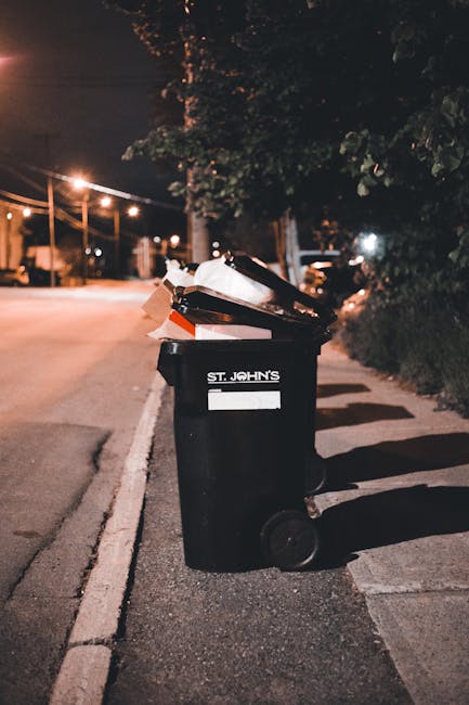 A sanitation worker in a green uniform and protective helmet is seen standing beside the rear of a garbage collection truck on a quiet street lined with trees. The truck, primarily white with red and 
