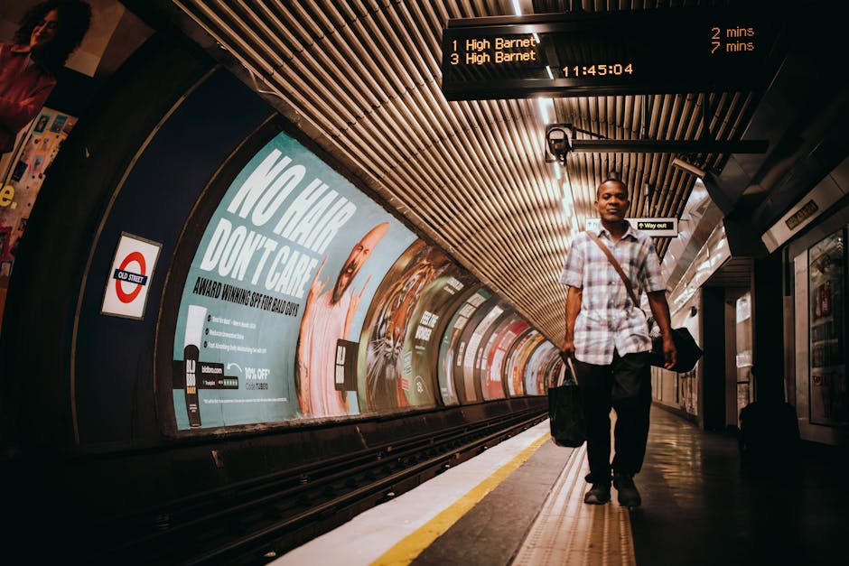 A man with dark skin, wearing a short-sleeved checked shirt and dark trousers, is walking along a train station platform carrying a black shoulder bag and a smaller black case in his left hand. Behind